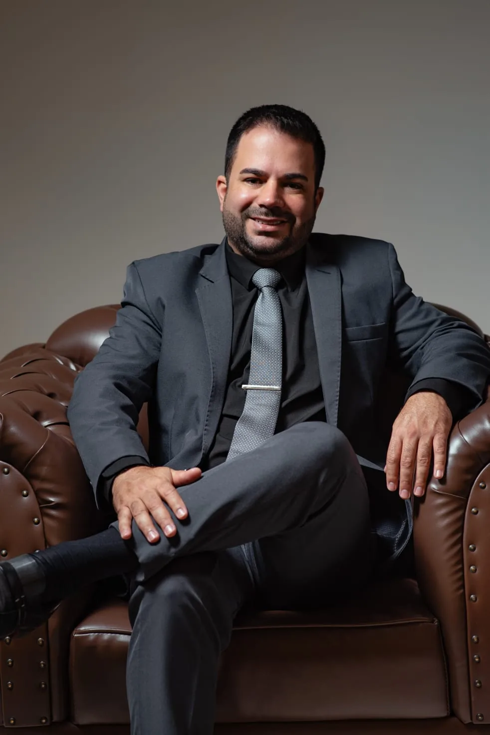 Michel Moreira sorrindo, sentado em uma poltrona chesterfield marrom